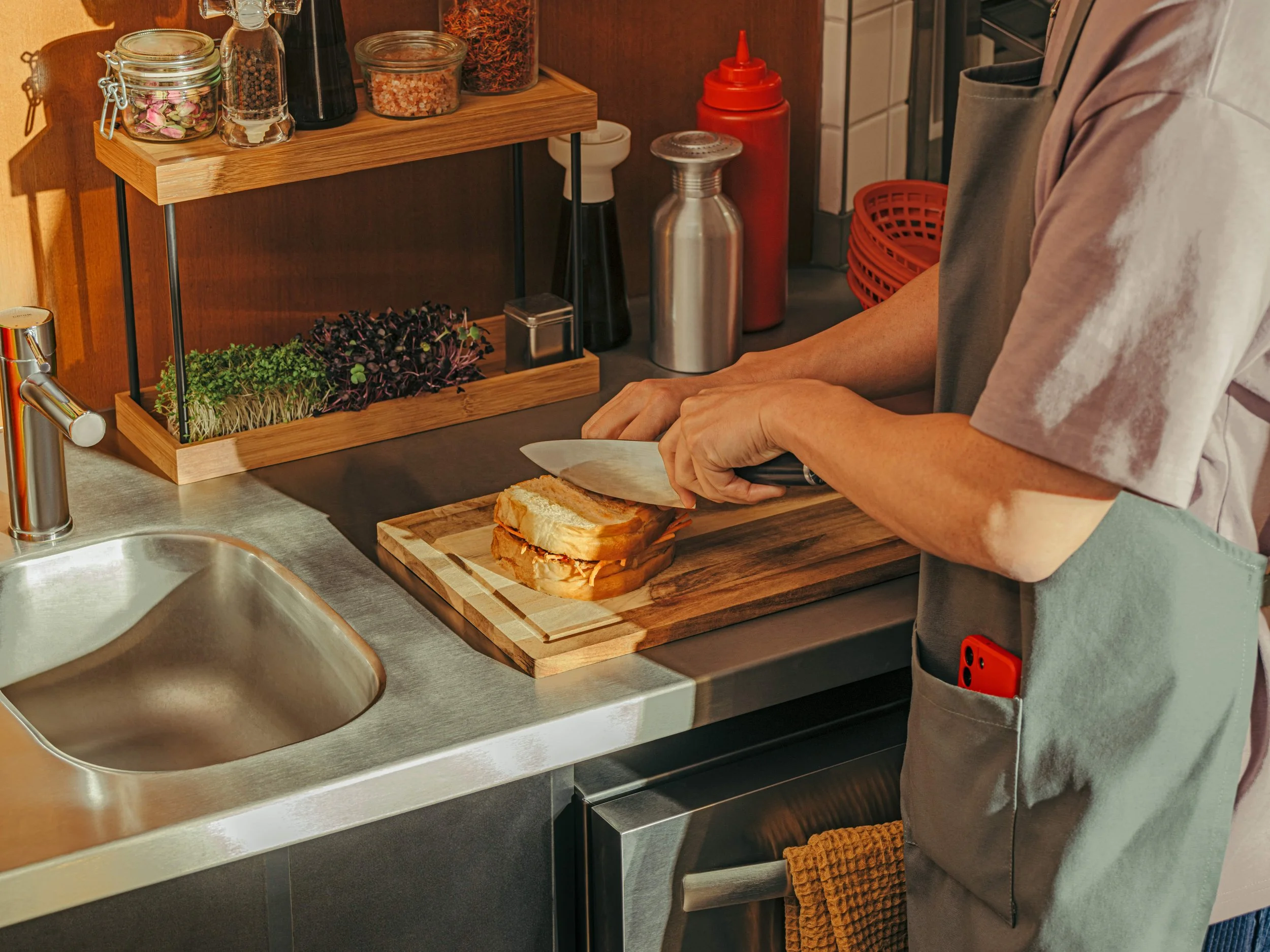 A person slicing a sandwich on a wooden cutting board in a modern kitchen, with various spices and herbs on a wooden shelf in the background.