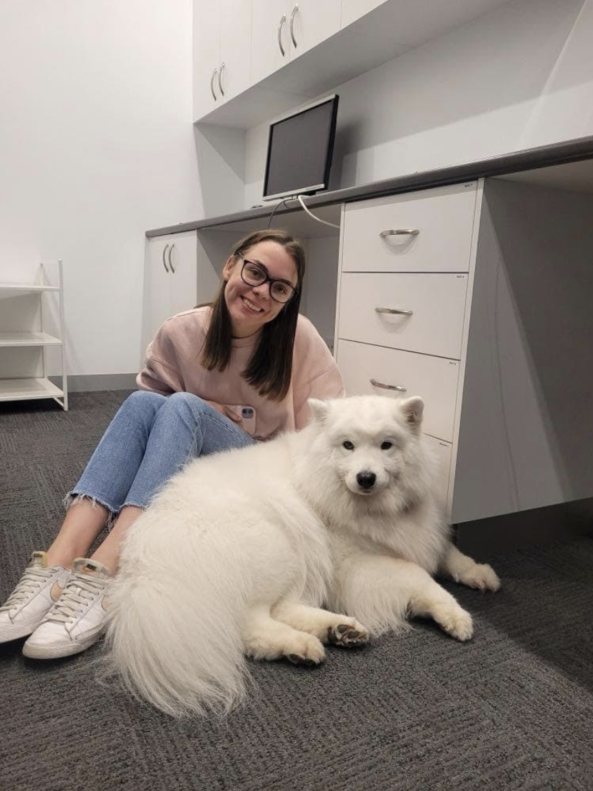 A woman with glasses, wearing a pink sweatshirt and blue jeans, sitting on the floor smiling at the camera next to a large white fluffy dog lying on the carpeted floor in an office or classroom setting.