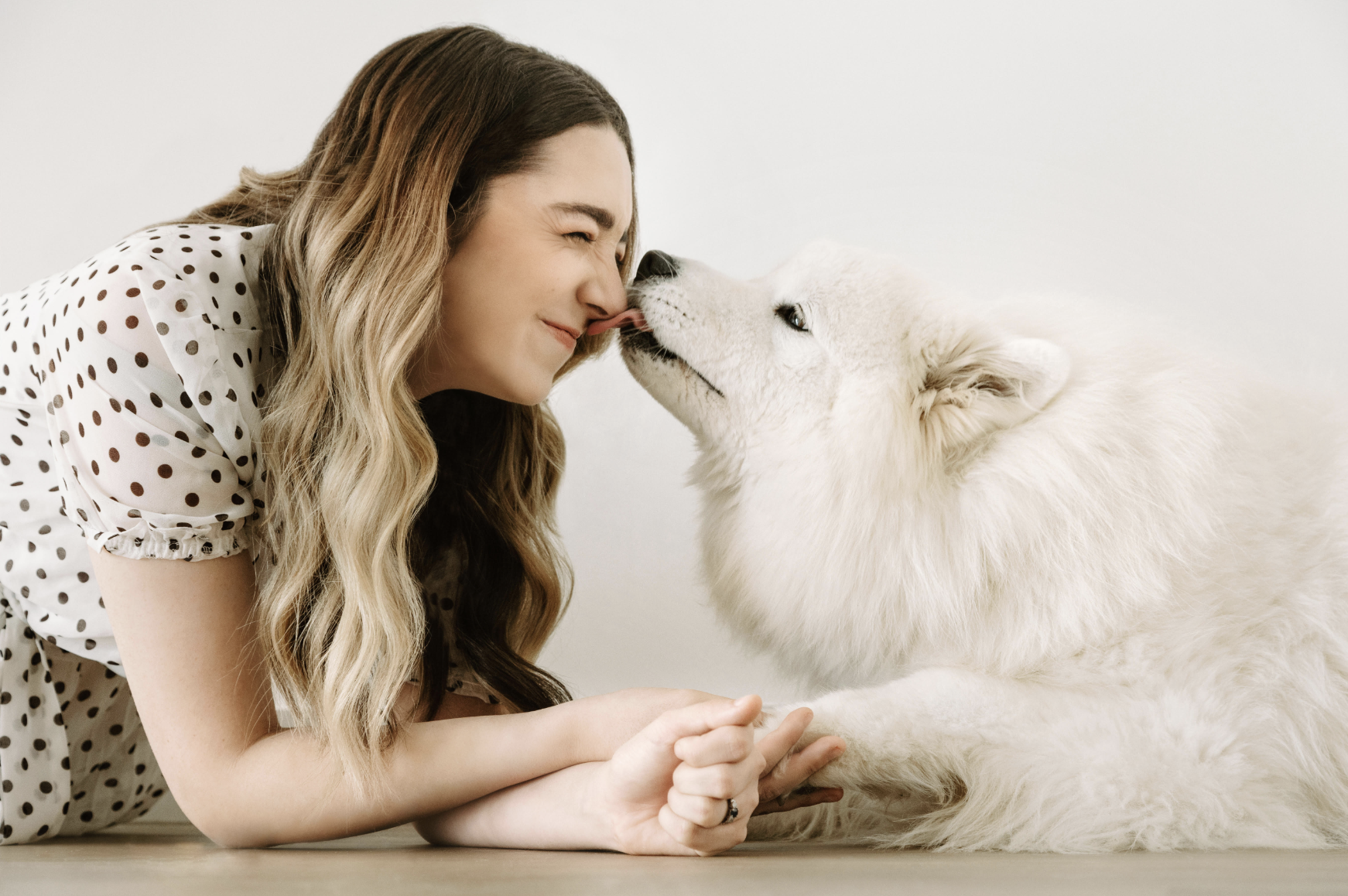 A woman with long wavy hair and a polka dot blouse smiling and leaning close to a white fluffy puppy, touching noses in a white room.