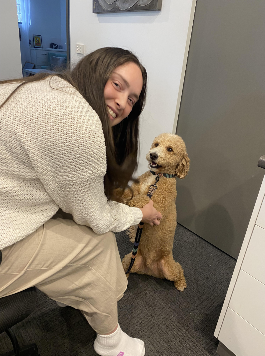 A woman with long brown hair, wearing a cream sweater and beige pants, kneeling and smiling while holding a tan curly-haired dog inside an office or room. The dog is standing on its hind legs with a collar and leash and looking at the camera.