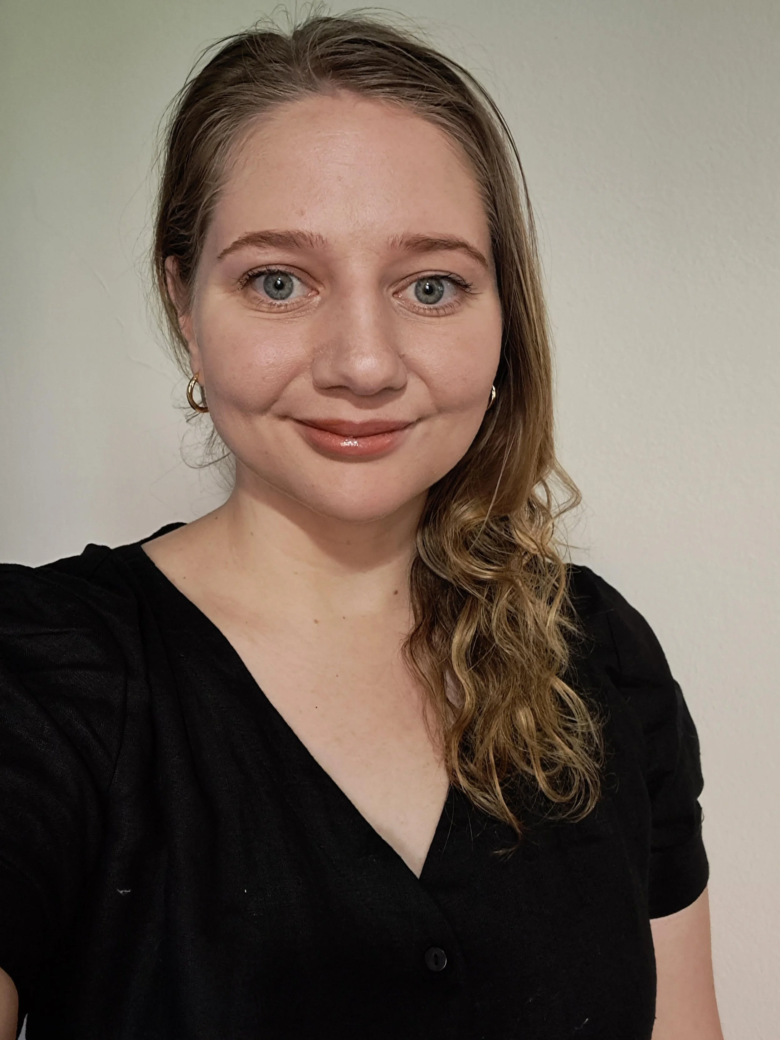 A young female psychologist with brown hair and light skin, wearing gold hoop earrings and a cream-colored sweater, looking directly at the camera with a neutral background.