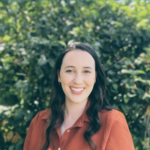 A young female speech pathologist with long dark hair smiling and standing outdoors in front of green foliage