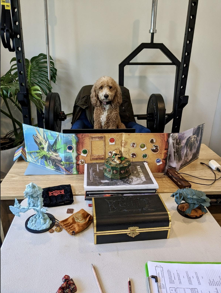 A dog sitting behind a table with gaming materials, a dice, and figurines, in front of a gaming setup, with a weightlifting rack, weights, and a plant in the background.