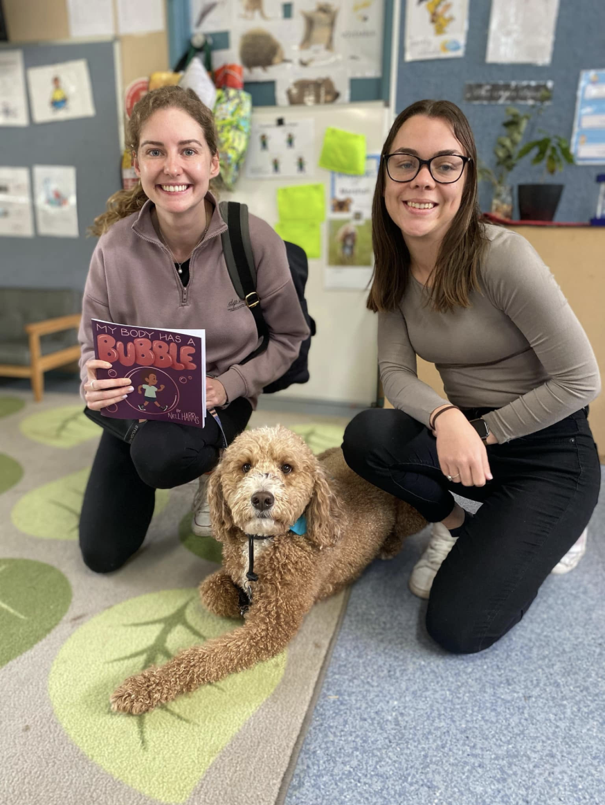 Two women kneeling on a leaf-patterned rug with a poodle dog between them in a classroom setting, one woman is holding a children's book titled 'My Body Has a Bubble' and both women are smiling.