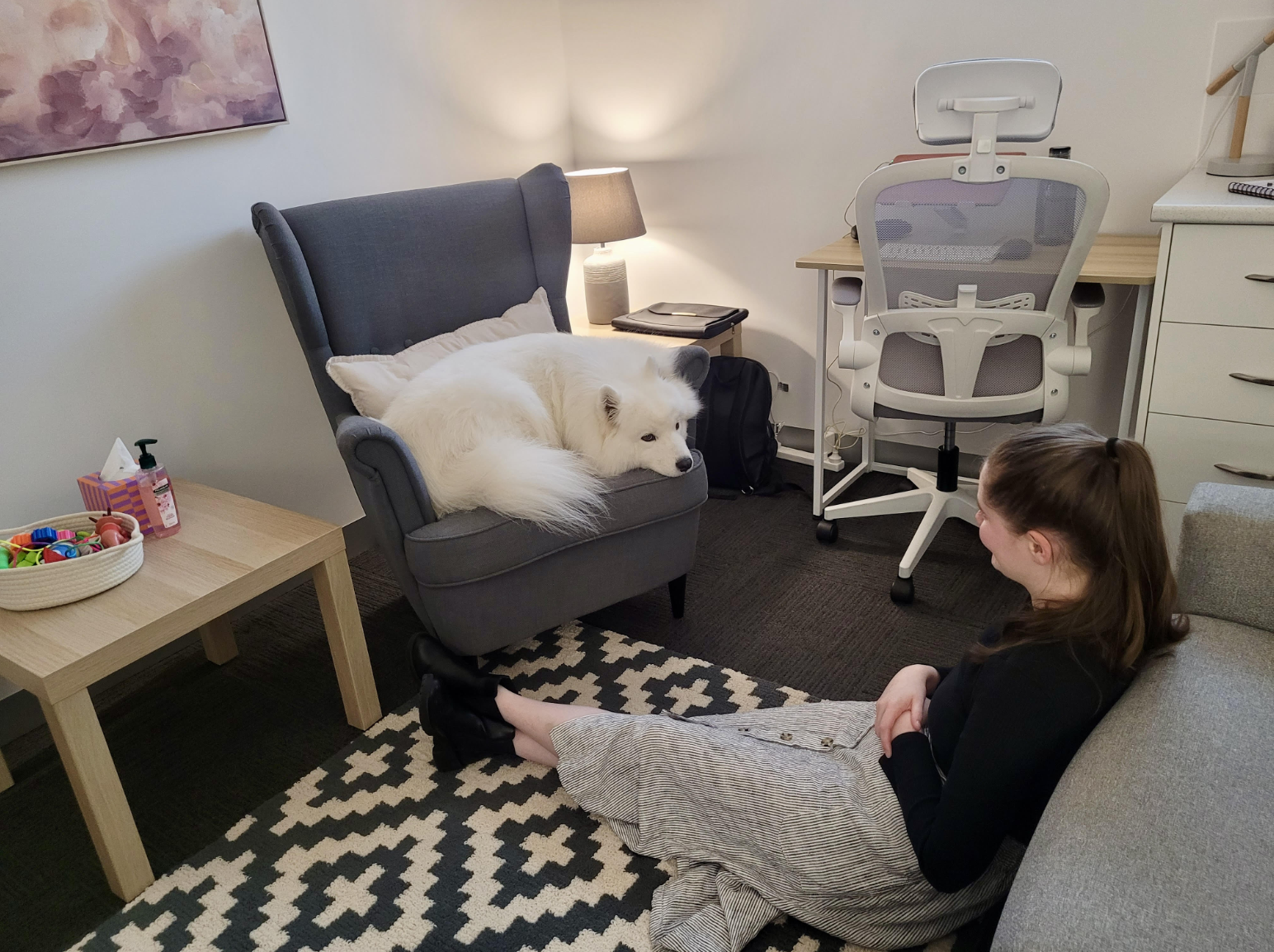 A woman sitting on the floor in front of a gray armchair with a large white dog lying on it. The room has a patterned black and white rug, a small wooden coffee table with toys and hand sanitizer, a white desk chair, and a desk in the background.
