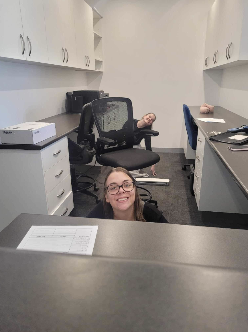 Two women in an office, one smiling at the camera from behind a desk, the other upside down and peeking from behind a chair, indoors with white cabinets and office furniture.