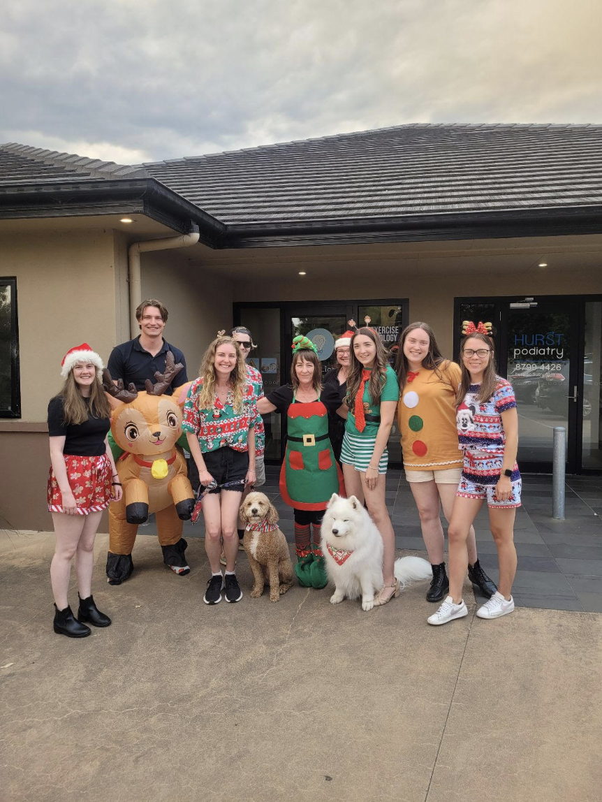 Group of people dressed in Christmas-themed outfits standing outside a building with holiday decorations, accompanied by dogs and inflatable reindeer and Santa.