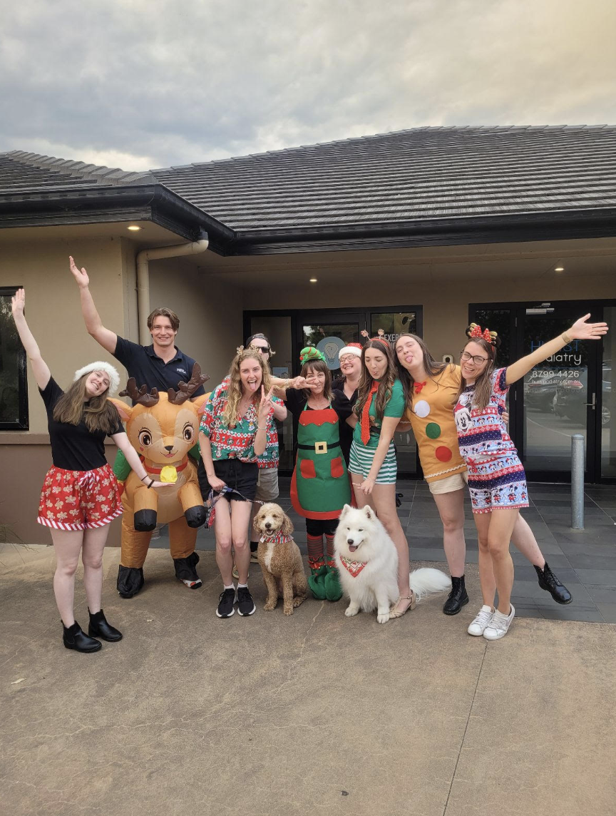 Group of people in festive holiday attire, including Christmas sweaters and accessories, standing outside a building with two dogs and a reindeer costume, smiling and posing for a photo.