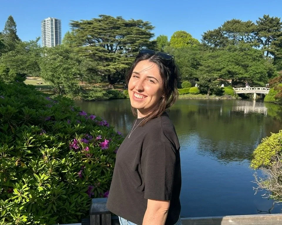A smiling young woman with dark hair, wearing a black top and sunglasses on her head, stands near a pond in a lush green park with trees and purple flowers, under a clear blue sky.