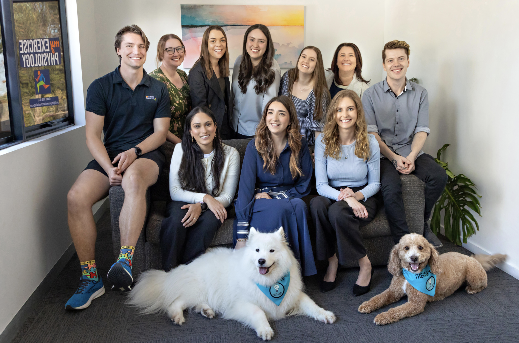 Group of ten people and two therapy dogs in an office. The people are smiling, seated and standing, with a colorful abstract painting on the wall behind them. The therapy dogs, one white and fluffy and the other small and curly-haired, are lying on t