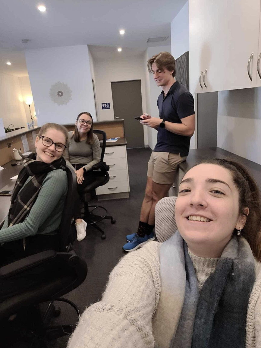 Four young adults in an office taking a selfie, smiling and casual. One woman in the foreground has a sweater and scarf, another woman in the background wears glasses and a gray sweater, a young man standing with a phone wears a navy shirt and beige