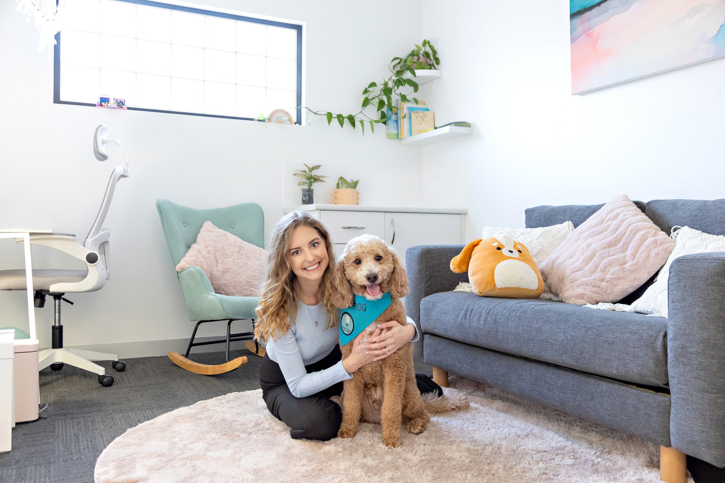 A woman sitting on a pink carpet in a modern living room, holding a light brown poodle wearing a blue bandana that says "Dog". The room has a gray sofa with pink and white pillows, a plush dog-shaped pillow, a light green rocking chair, and various w