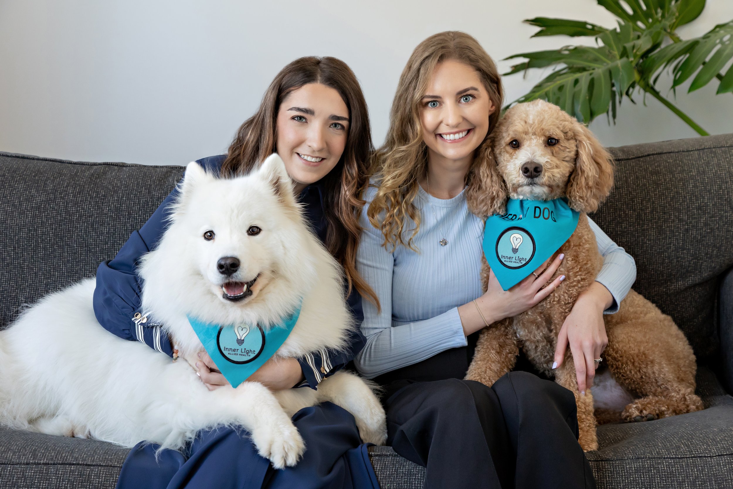 Two women sitting on a gray couch holding a white fluffy dog and a brown curly-haired dog, both wearing blue bandanas with a lightbulb and the words "Inner Light" on them, smiling at the camera, with a green plant in the background.