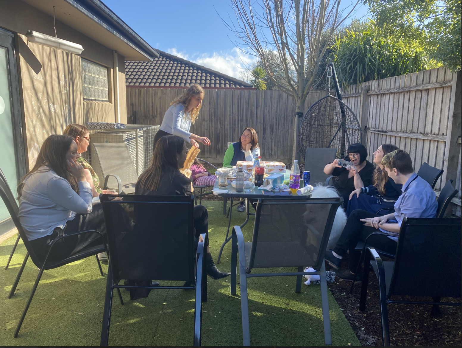 Group of women celebrating birthday outdoors in a backyard, sitting around a table with food and drinks, with one woman standing and giving a gift, and others enjoying the moment.