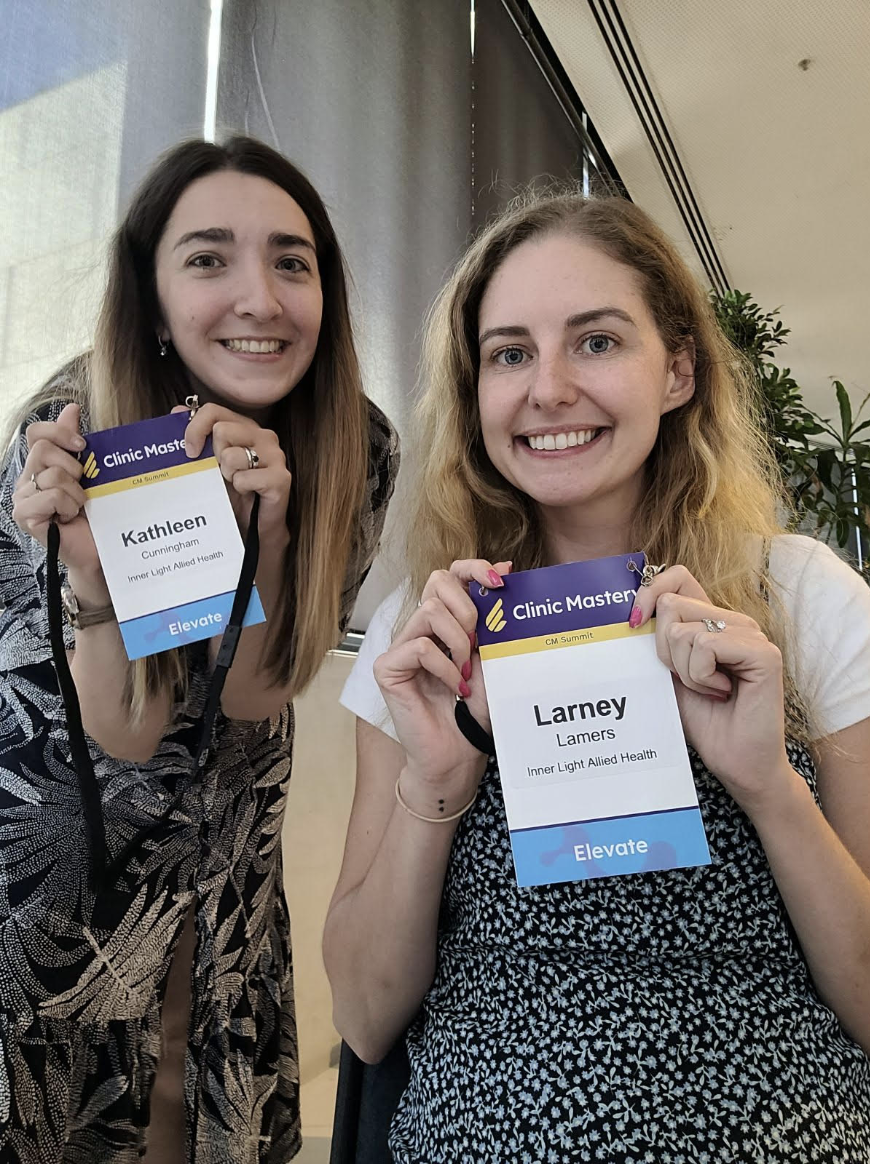 Two women smiling, holding conference name tags with 'Elevate' written at the bottom, in a brightly lit room with a plant in the background.