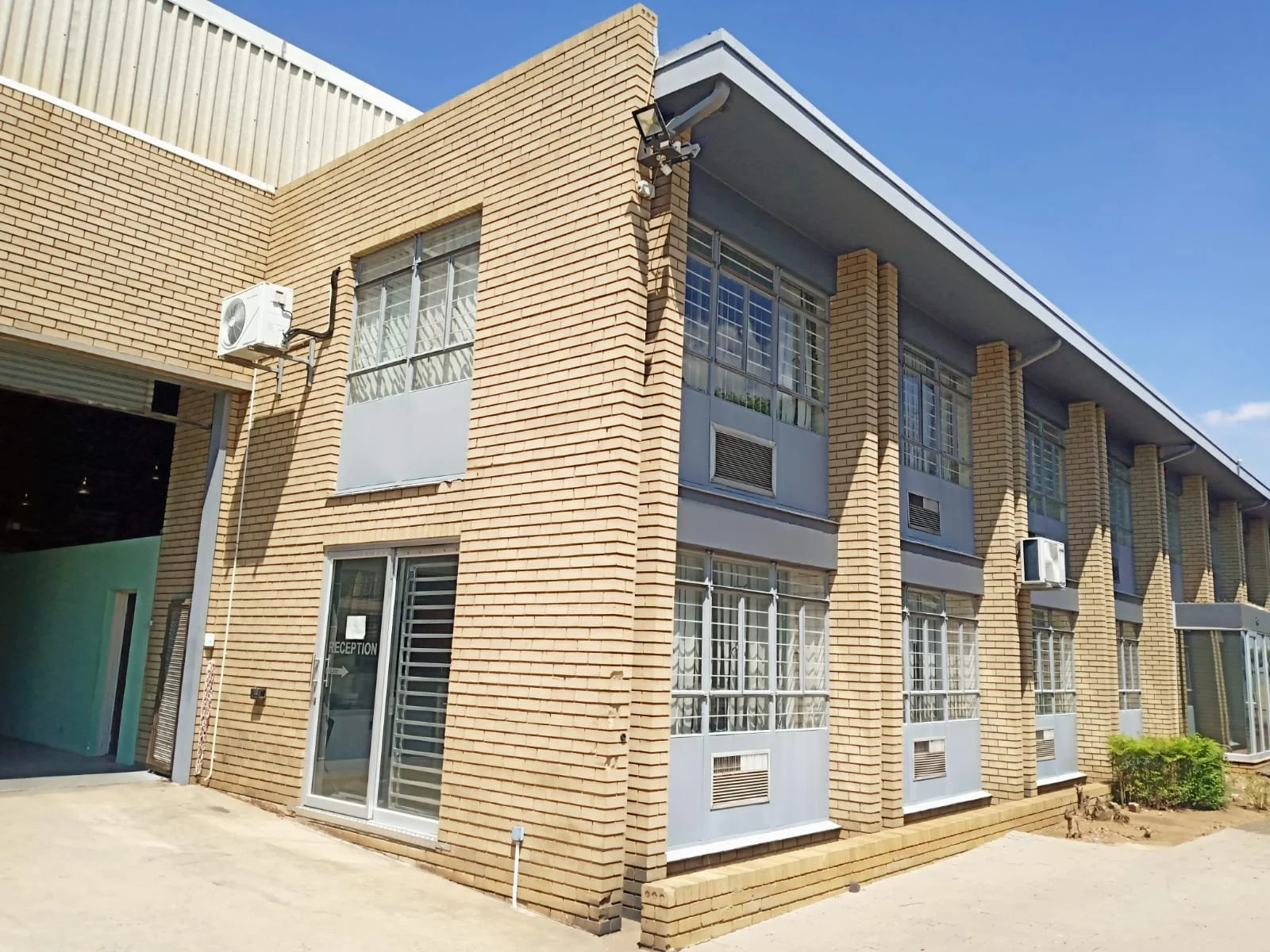 A two-story brick building with multiple large windows and air conditioning units mounted outside, under a clear blue sky.