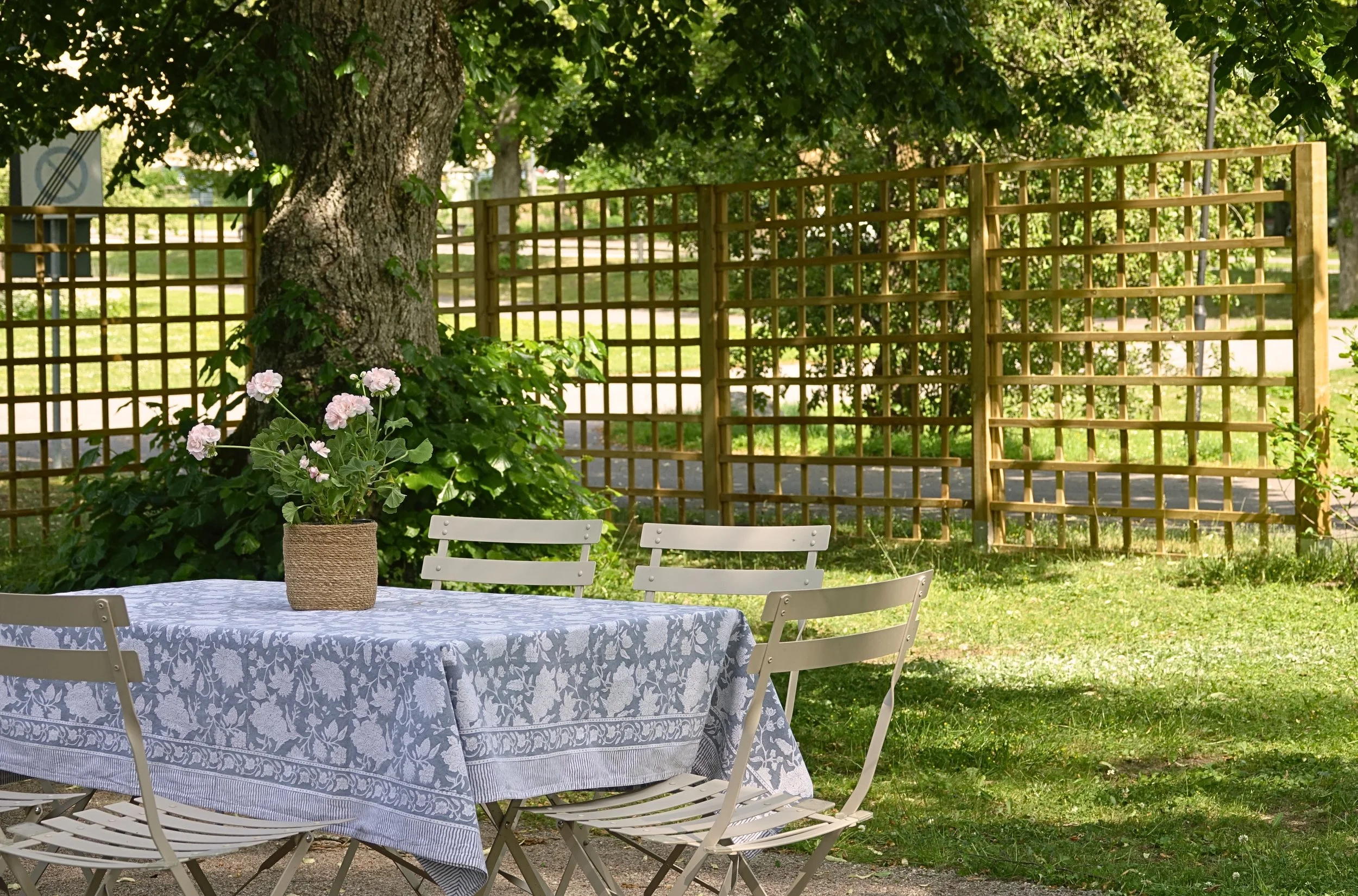 En lekplats eller trädgård med ett runt bord täckt av ett blåvitt blomsterfärgat tyg och fyra vita stolar. En krukväxt med rosa blommor står på bordet. Det finns en stor träd och en trästaket i bakgrunden.