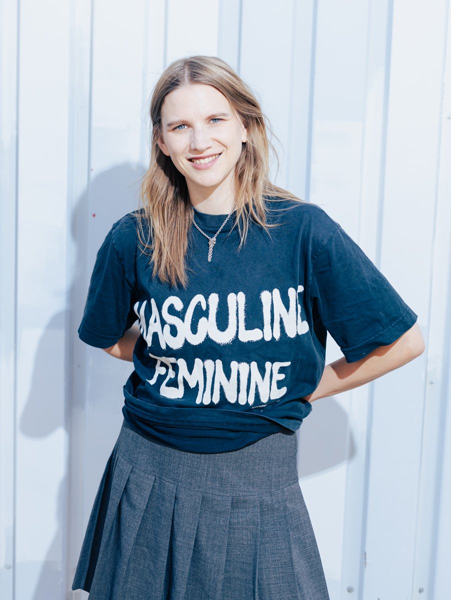 Foto von Carla-Frieda Nettelnbreker ; steht vor einer weißen Wand, trägt ein dunkles T-Shirt mit weißen Schriftzug "MÄNNLICH WEIBLICH"