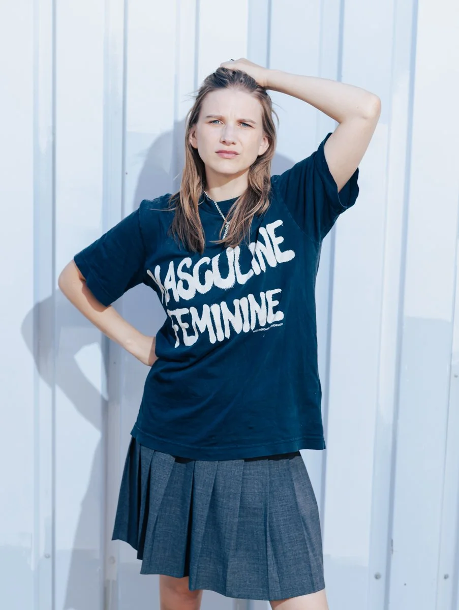 Foto von Carla-Frieda Nettelnbreker,  steht vor einer weißen Wand, trägt ein T-Shirt mit dem Text 'HASSULINE FEMININE' und schaut nachdenklich, während sie den Kopf mit einer Hand hält.