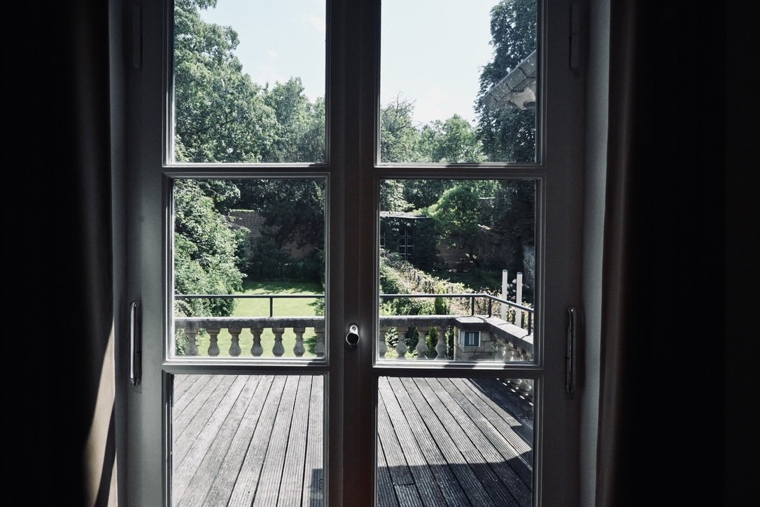 View through a glass door showing a wooden deck and a lush green garden with trees and shrubs.