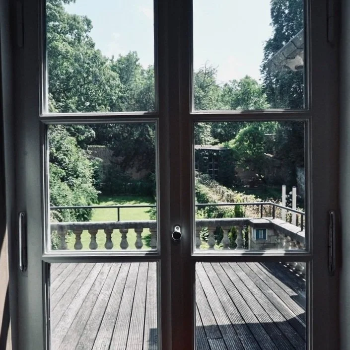View through glass doors onto a wooden deck and lush green backyard with trees and a small structure in the distance.