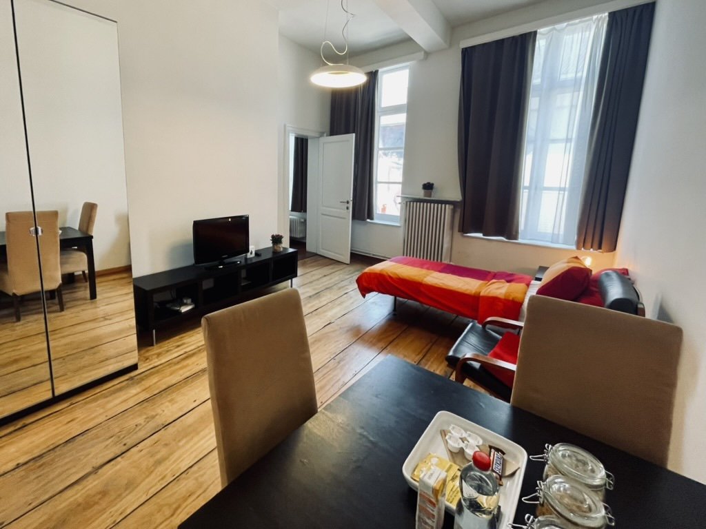 Living room with a single bed with red and orange bedding, a black sofa, a TV on a black stand, a dining table with chairs, and large windows with dark curtains.