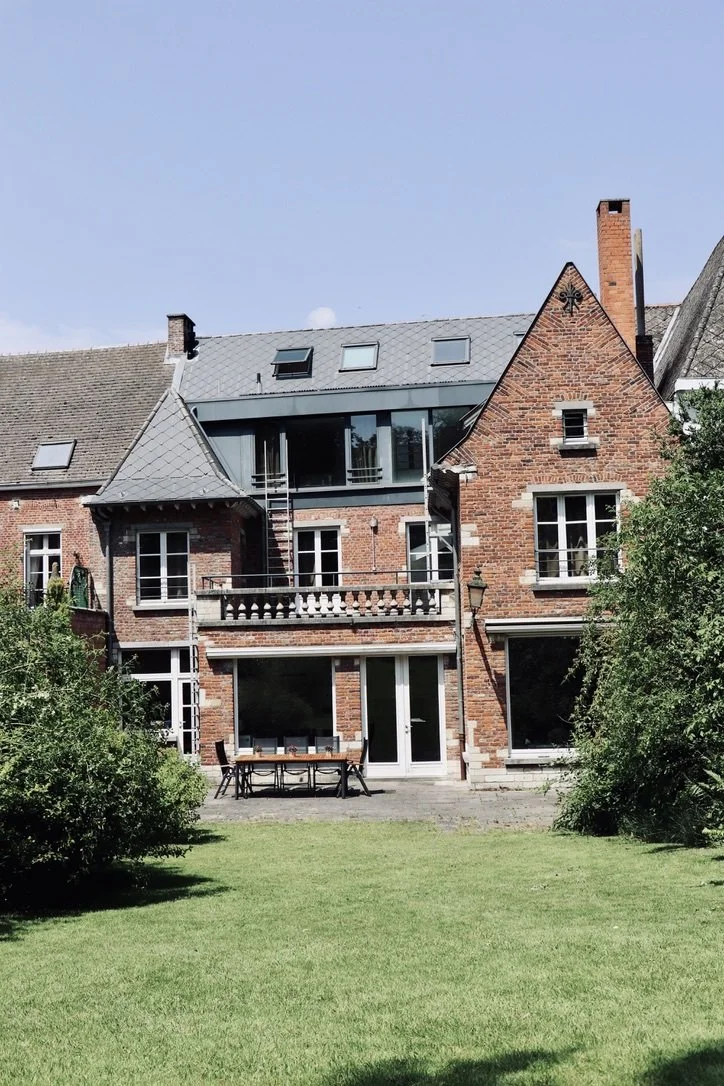 Brick residential house with a backyard patio, surrounded by bushes and grass, under a clear blue sky.