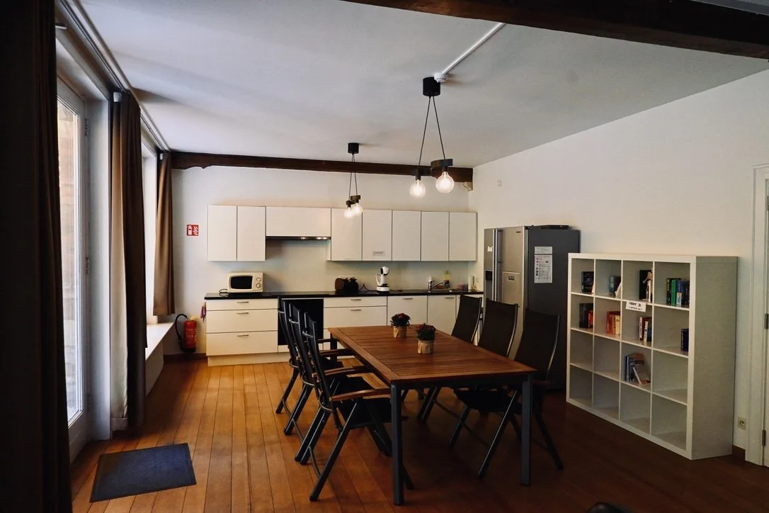 Kitchen and dining area with white cabinets, a microwave, refrigerator, and a wooden dining table with eight chairs in a room with hardwood floors and large windows.