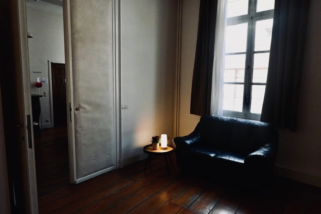 A cozy room corner with a black leather couch, a small round side table with a lamp and a potted plant, a window with curtains, and a wooden floor.
