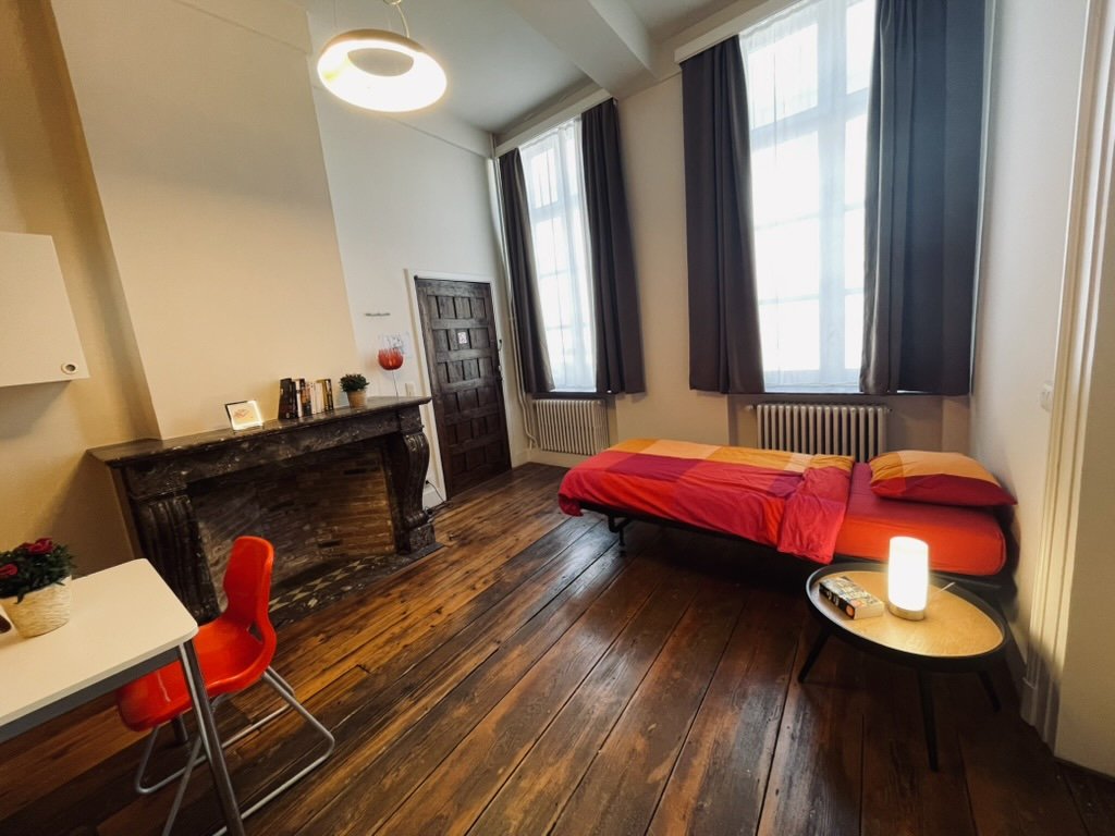 Small bedroom with hardwood floors, a bed with orange and red bedding, a white desk with a red chair, a black side table with a lamp, large windows with dark curtains, a fireplace mantle, and a ceiling light.