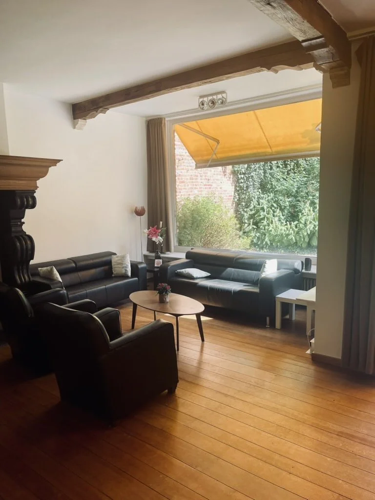 Living room with black leather sofas, a wooden coffee table with a plant, armchair, tall lamp, large window with beige curtains, and wooden floor.
