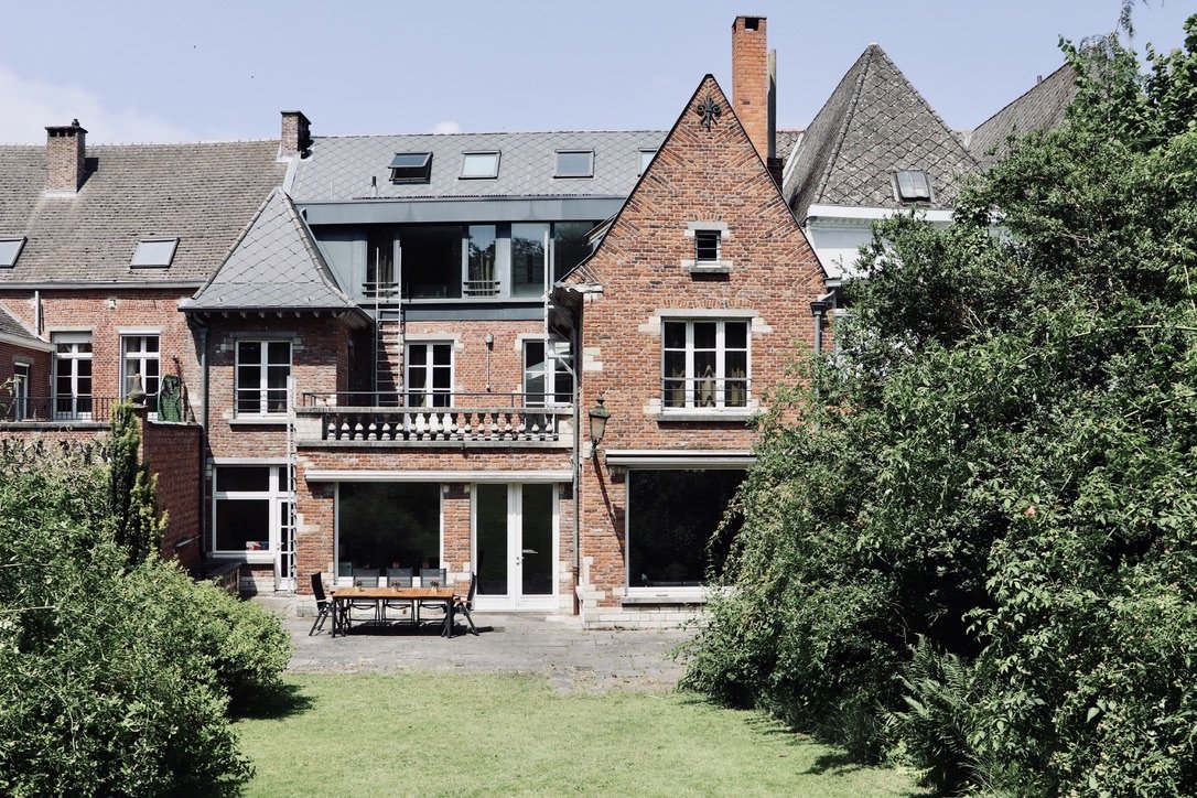 Rear view of a multi-story brick house with modern and traditional architectural elements, surrounded by greenery and a lawn with outdoor furniture.