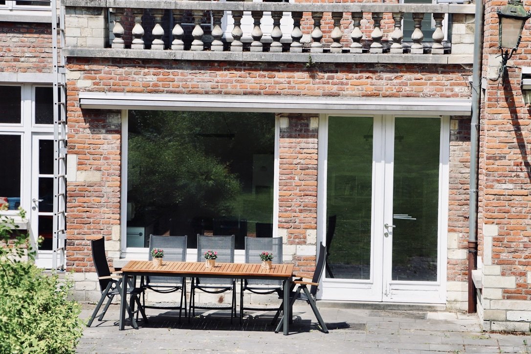 Backyard patio with a wooden dining table, six chairs, and small potted plants, in front of a brick house with large glass doors and windows.