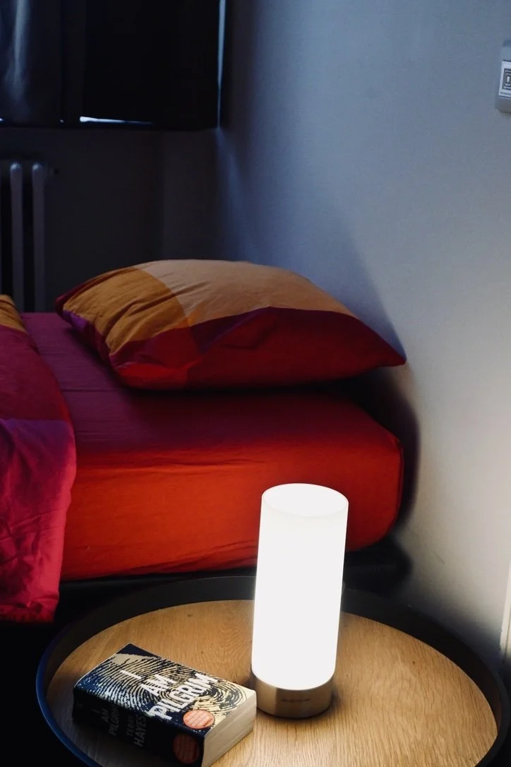 A bedside table with a white cylindrical lamp and a paperback book next to a bed with red and orange bedding and a pillow, in a dimly lit room.