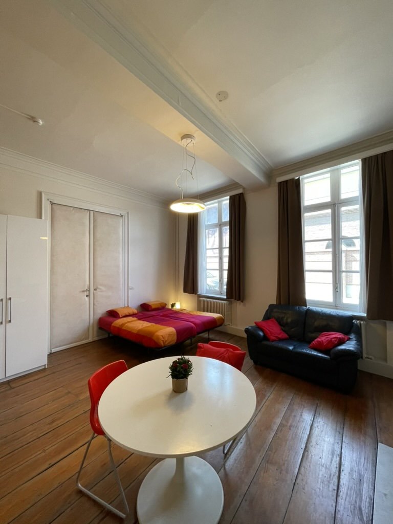 A living room with a double bed, a black couch with red pillows, a round white table with a small potted plant, large windows with brown curtains, and wooden floors.