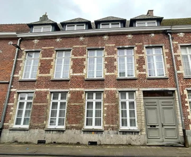 Three-story brick building with multiple white-framed windows and a large gray door, with a row of small attic windows on the roof and a cloudy sky above.