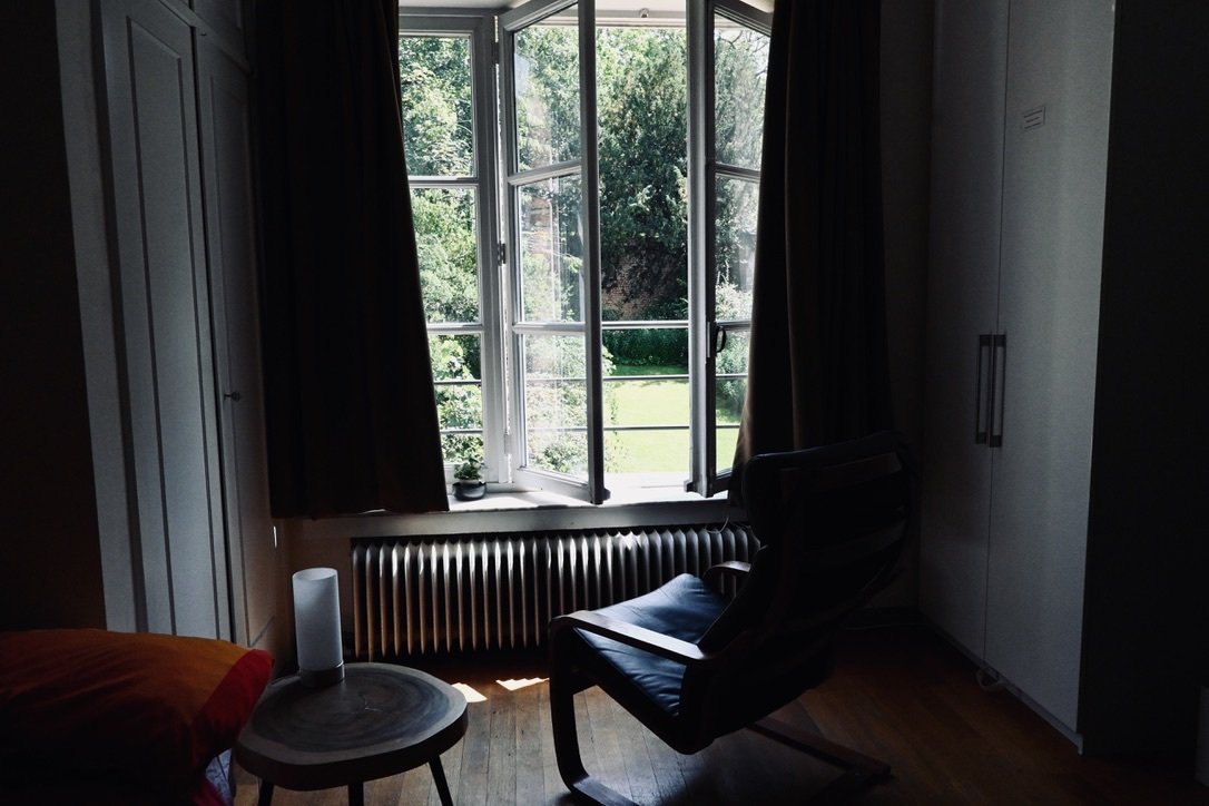 A bedroom with an open window revealing trees outside, a black chair, a small table with a white object on it, a bed with a red blanket, and a white wardrobe.