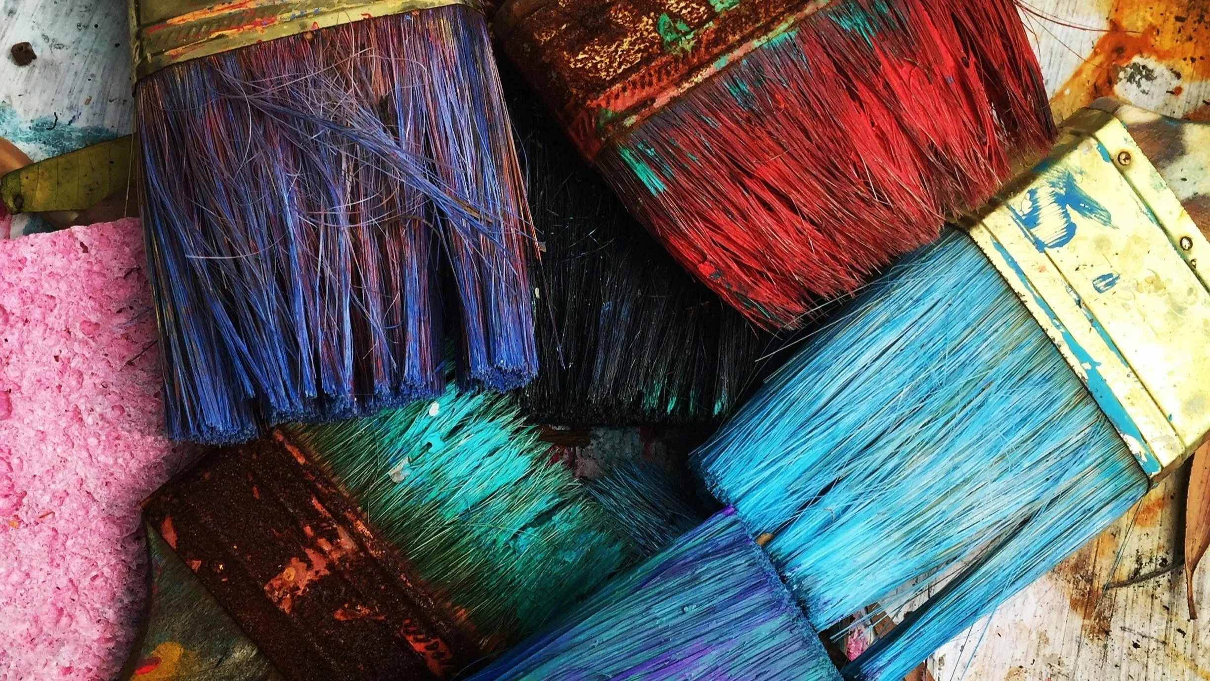 Close-up of multiple paintbrushes with colorful paint on bristles and handles, arranged on a table with paint splatters.