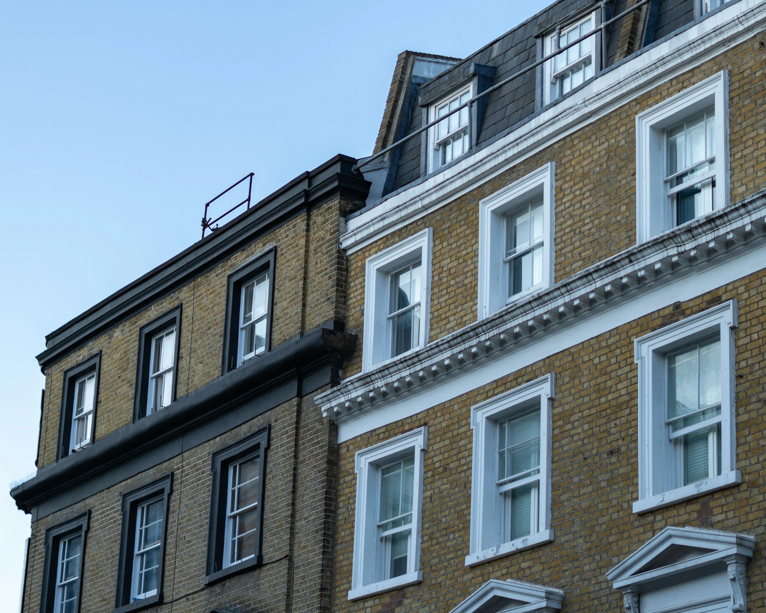Close-up of a multi-story building with yellow brick exterior, white window frames, and decorative white trim under the windows, with a portion of a black roof visible against a clear blue sky.