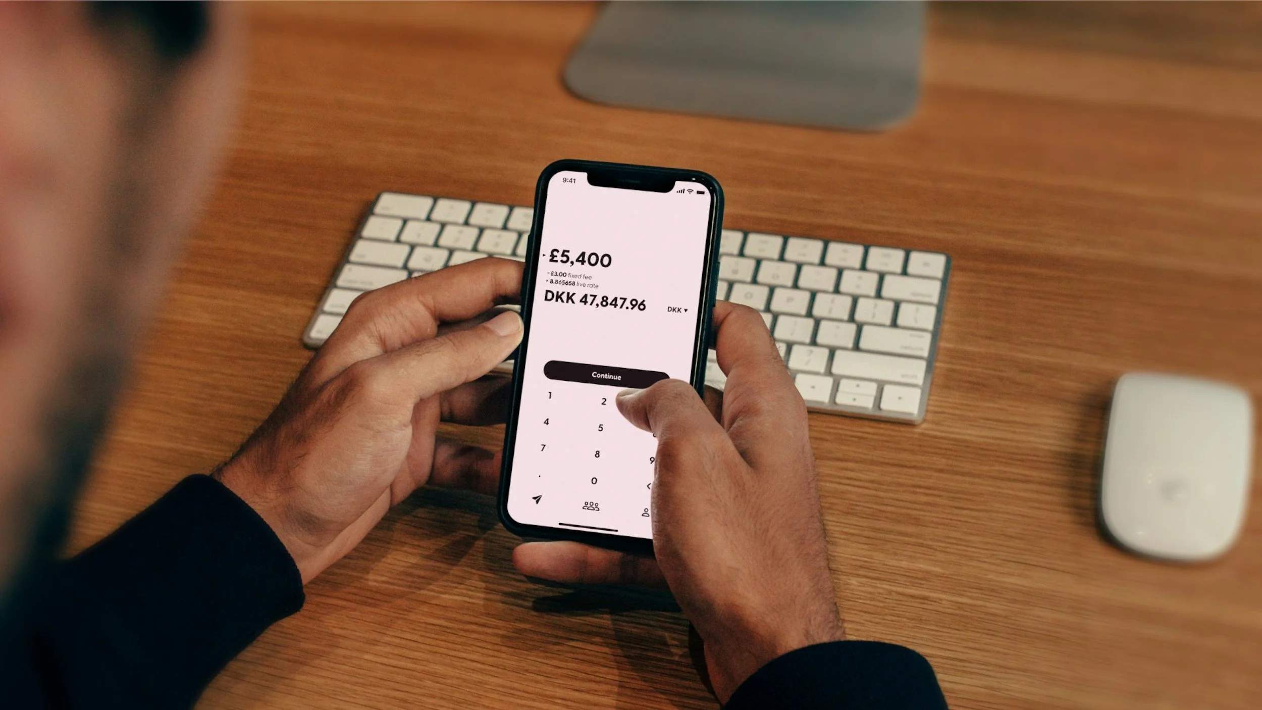 Person holding a smartphone displaying a financial app with text showing an amount of £5,400 and DKK 47,847.96 on a wooden desk with a wireless keyboard, mouse, and a monitor in the background.