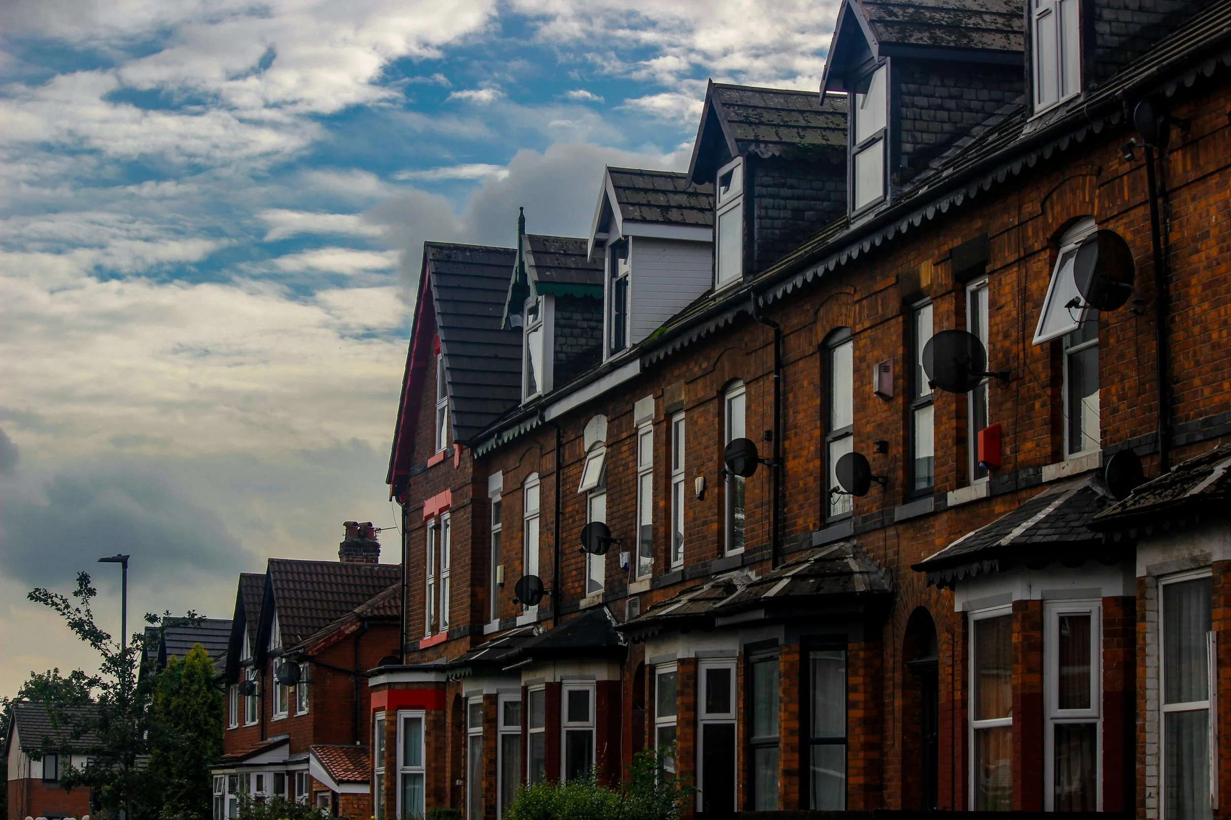 A row of brick apartment buildings with multiple windows, satellite dishes, and rooftops under a cloudy sky.