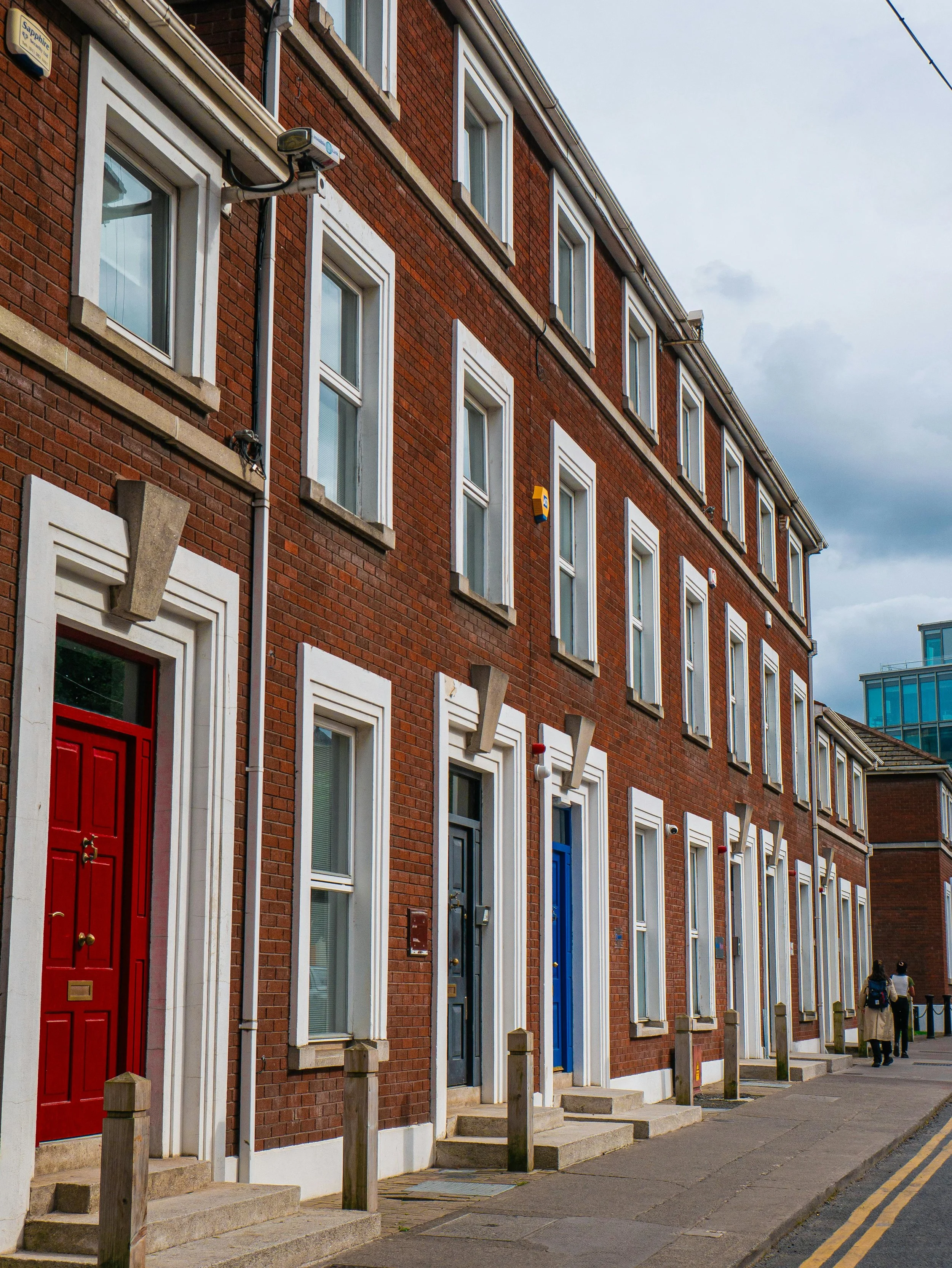 Red brick townhouses with colorful doors on a city street, cloudy sky in the background.