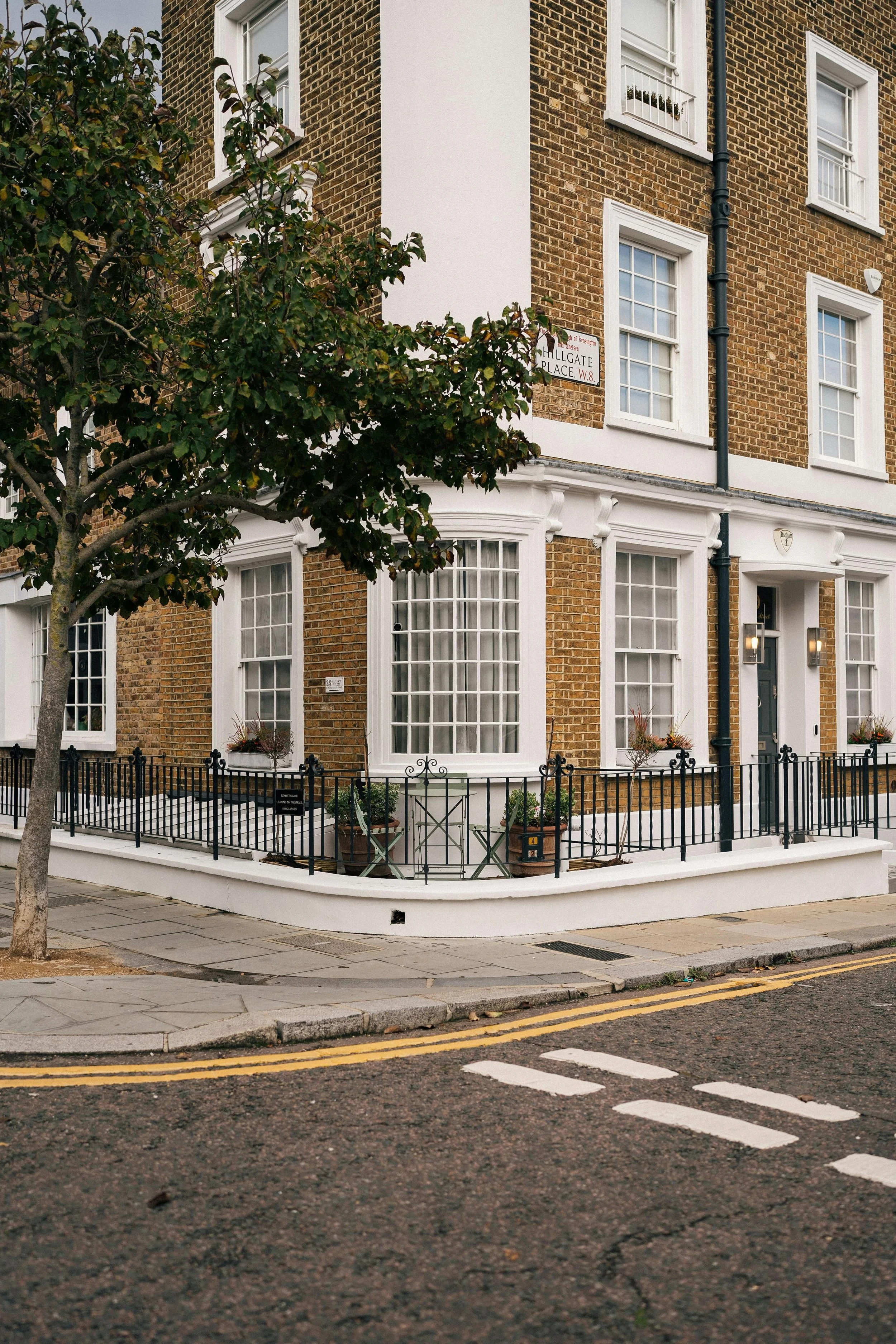 A corner of a brick building with large white-trimmed windows and black wrought-iron fencing, situated on a sidewalk with a tree in the foreground.