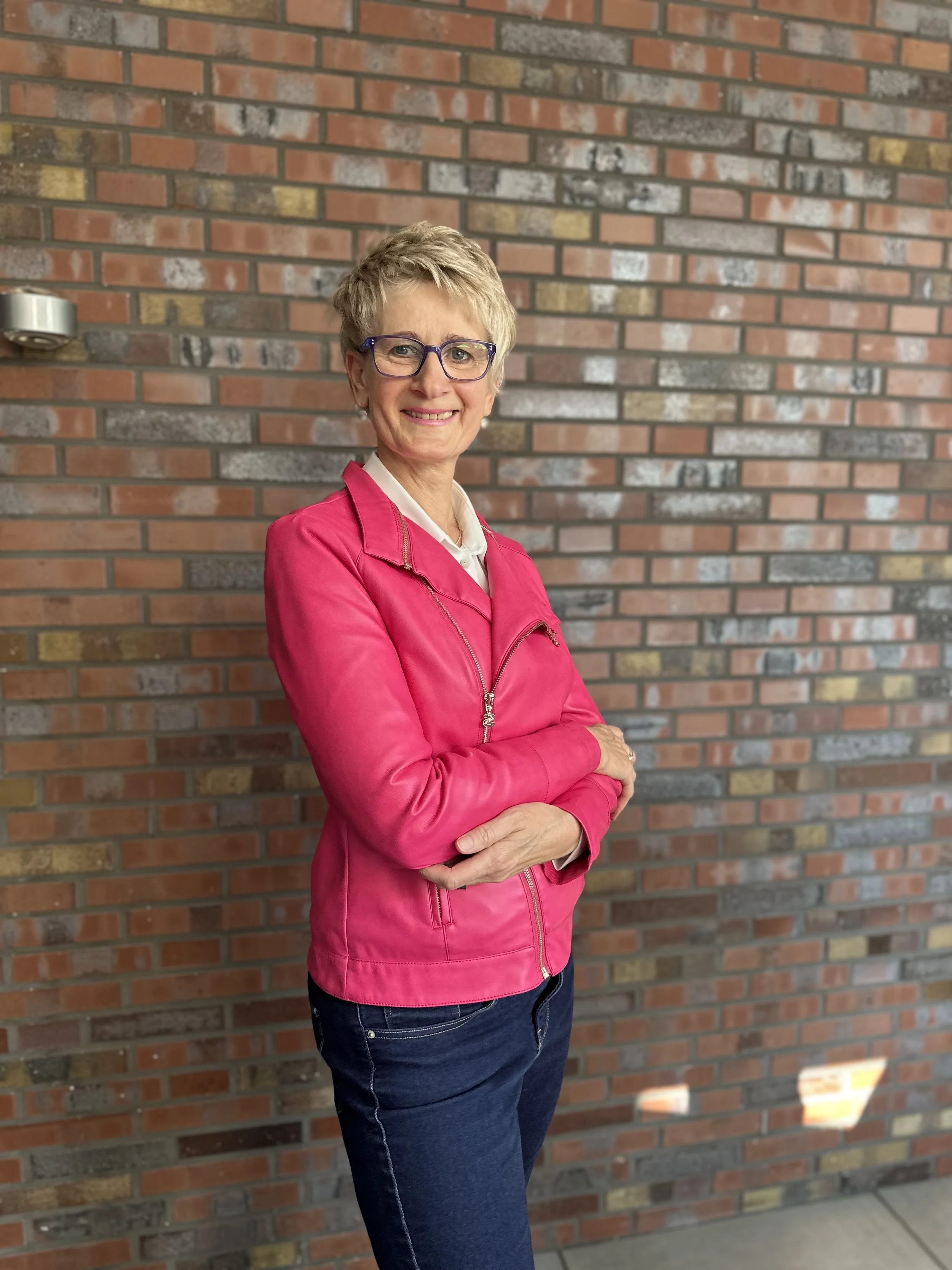 Eine ältere Frau mit kurzen grauen Haaren, Brille und einem freundlichen Lächeln steht vor einer Ziegelmauer, trägt eine pinke Jacke, einen weißen Hemdkragen und dunkle Jeans.