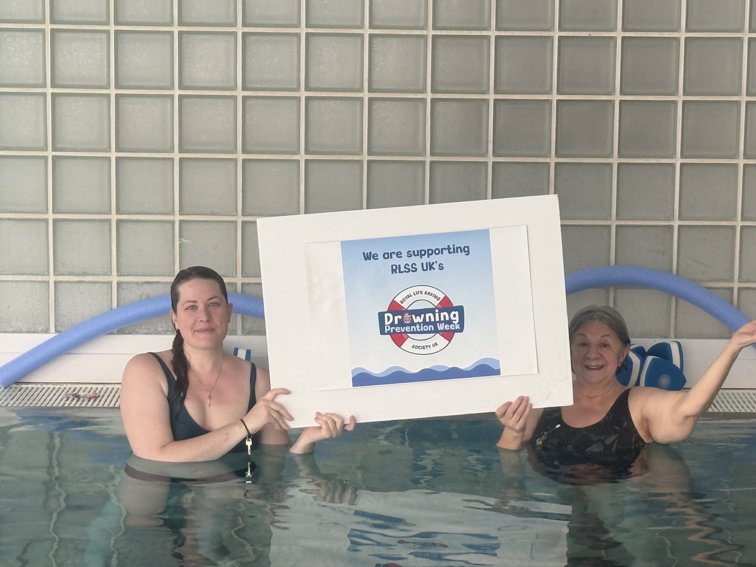 Two women in a swimming pool holding a sign supporting RLSS UK's drowning prevention week. One woman is on the left with dark hair in a braid, wearing a dark swimsuit. The other woman is on the right with short gray hair, wearing a dark top, smiling and holding part of the sign. The sign has the RLSS UK logo and the text 'We are supporting RLSS UK's Drowning Prevention Week'.