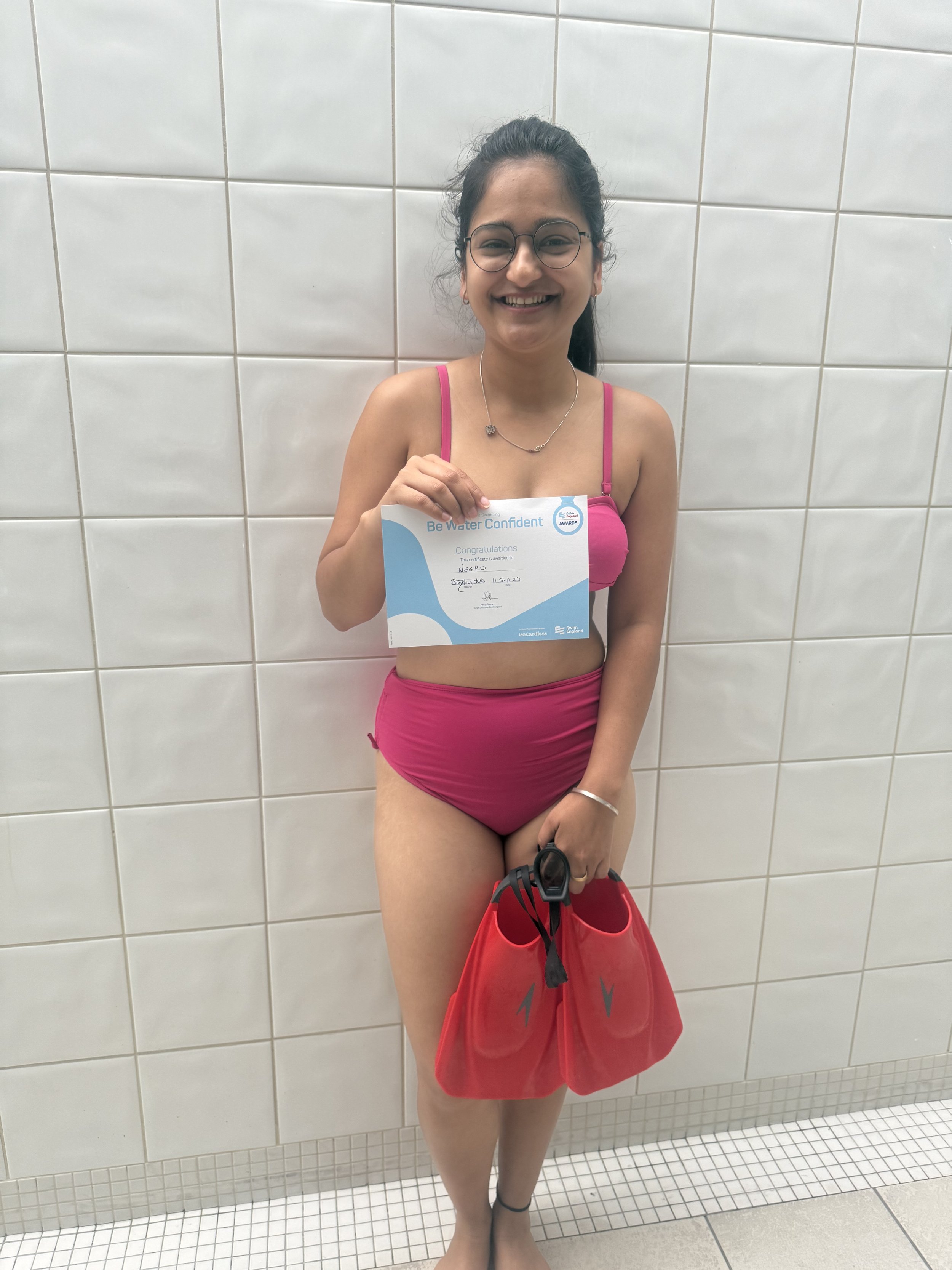 Young woman in pink swimwear, smiling, holding a water confidence certificate and red swim gear, standing against a tiled wall in a swimming pool area.
