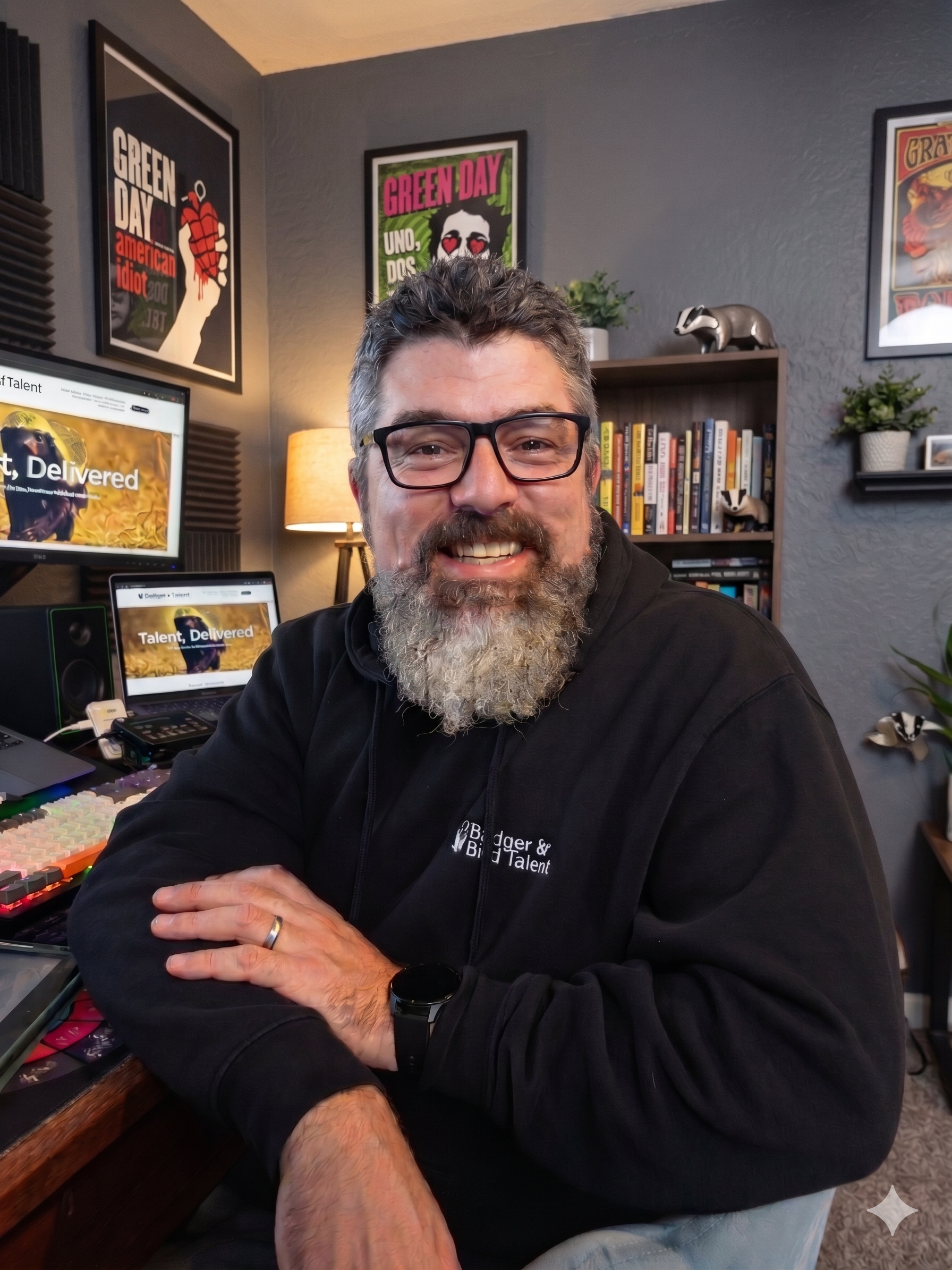 A smiling man with glasses and a beard, wearing a black sweatshirt, sitting at a desk with computer screens and audio equipment in a home office. The wall behind him has framed posters, including a Green Day album cover, and books on a shelf. There a