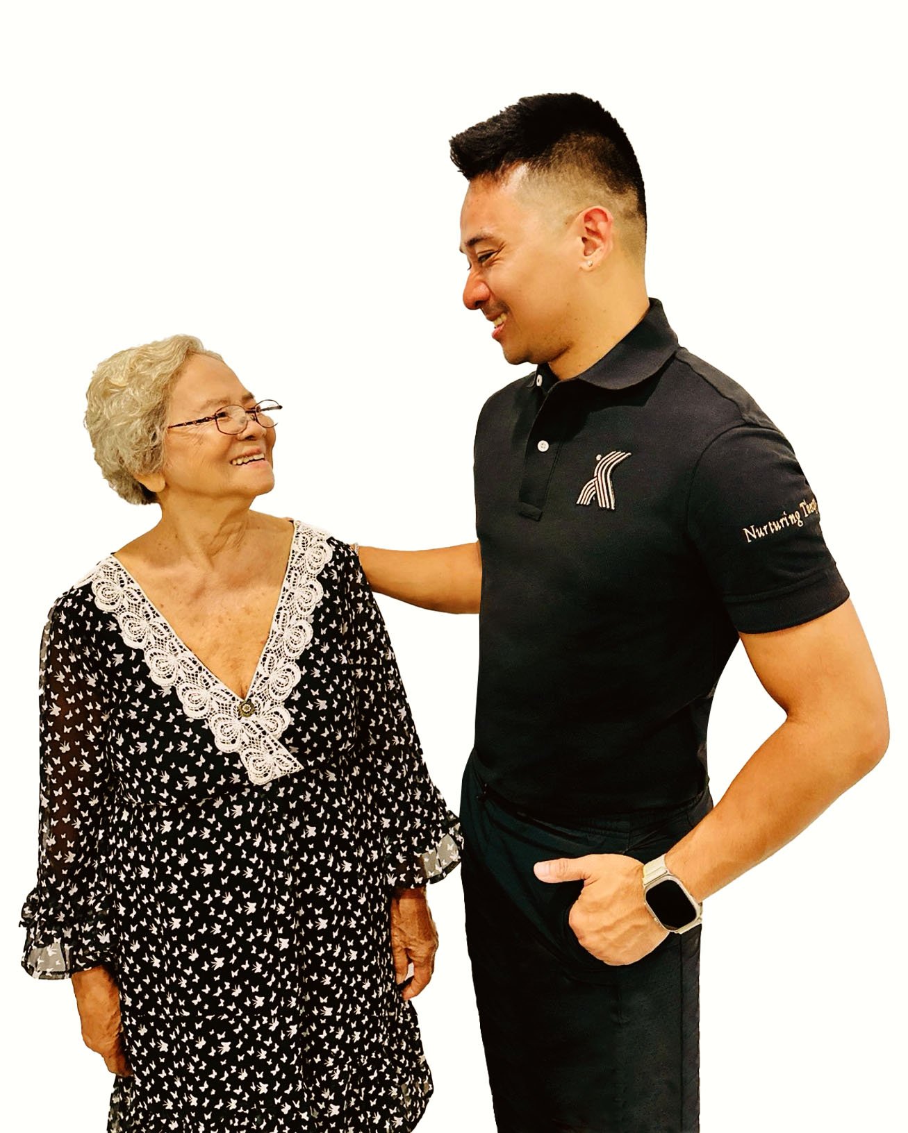An elderly woman and a young man smiling at each other. The elderly woman wears glasses and a black dress with white patterns and lace details. The young man wears a black polo shirt with a logo and has his arm around the elderly woman.