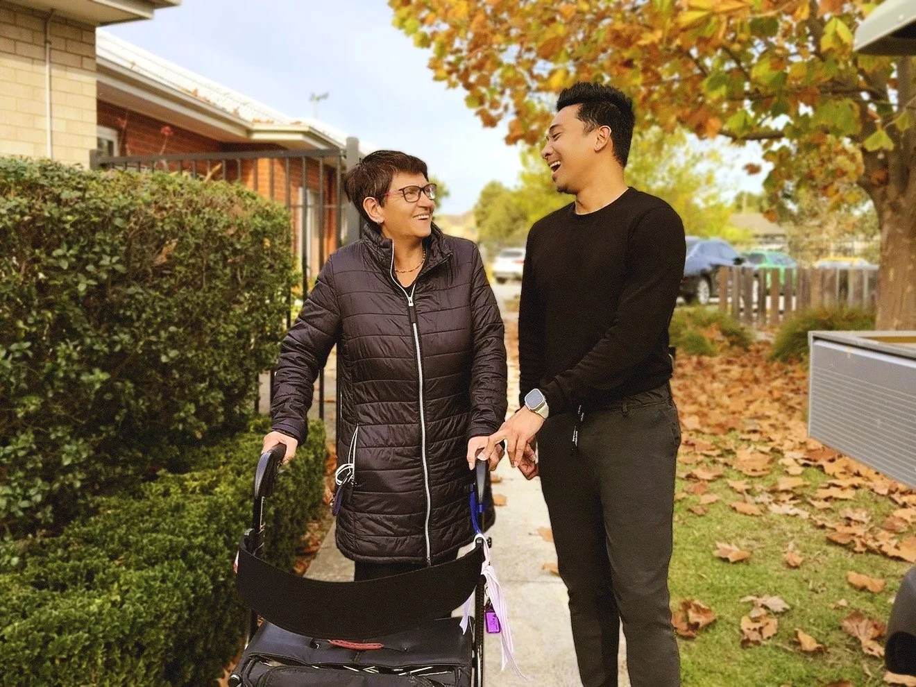 A woman with glasses and a dark jacket smiling and holding a walker, talking to a young man in a black shirt, both smiling and walking on a sidewalk with fallen leaves, autumn trees, houses, and parked cars in the background.