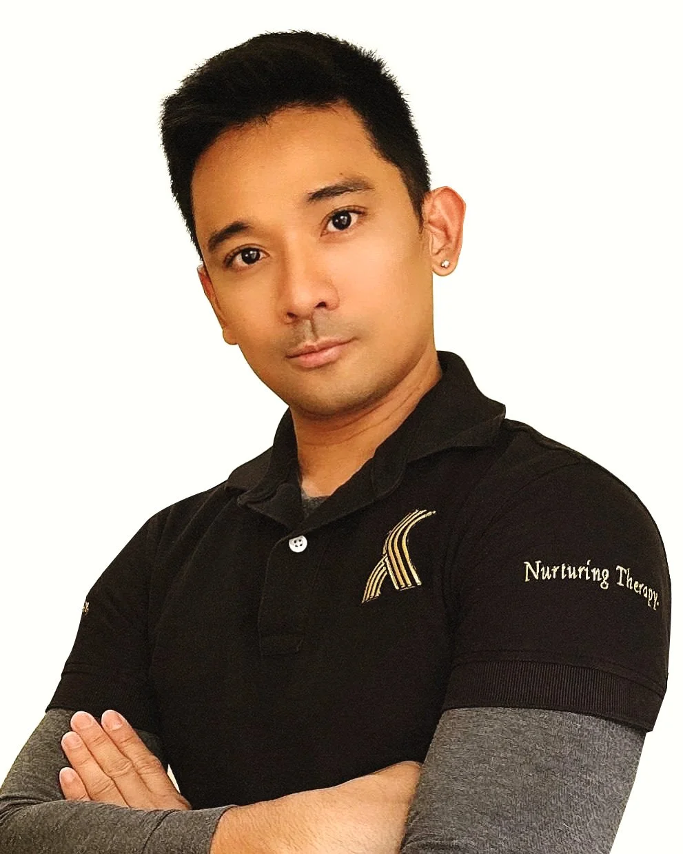 Young man with short black hair and a medium tan skin tone, wearing a black polo shirt with the KR Physiotherapy logo embroidered, paired with grey long sleeves underneath, arms crossed, and looking at the camera against a plain white background.